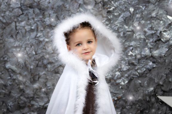 Navidad 2023 Niña pequeña con capa blanca y capucha, sonriendo frente a un fondo brillante.