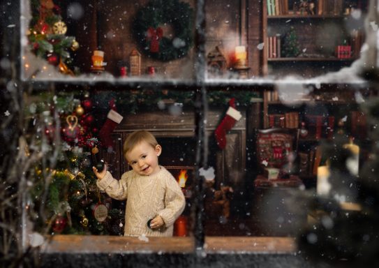 Navidad 2024 Niño pequeño mirando a través de una ventana mientras neva, decoraciones navideñas alrededor.