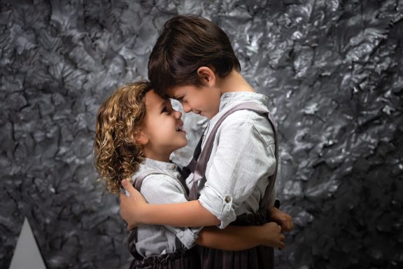 Navidad 2023 Dos niños abrazándose y sonriendo, mirándose a los ojos en un fondo gris.