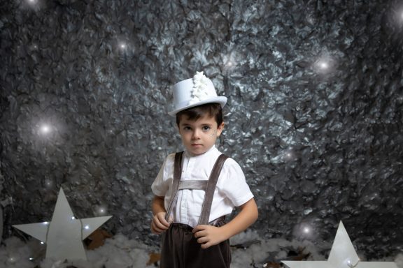 Navidad 2023 Niño con gorro blanco y tirantes, posando frente a fondo plateado decorado con estrellas.