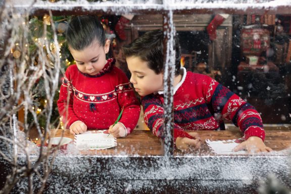 Navidad 2024 Niños escribiendo cartas de Navidad junto a una ventana decorada con nieve.
