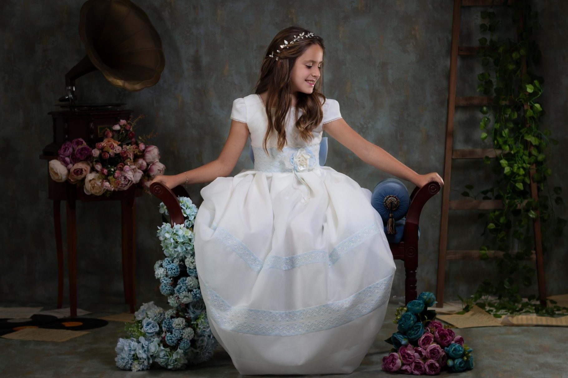 Niña sentada con un vestido blanco y azul, decorada con flores y una tiara.
