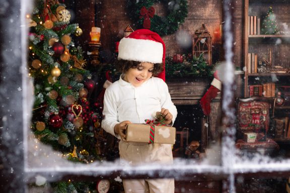 Navidad 2024 Niño con gorro de Navidad sosteniendo un regalo frente a un árbol decorado.